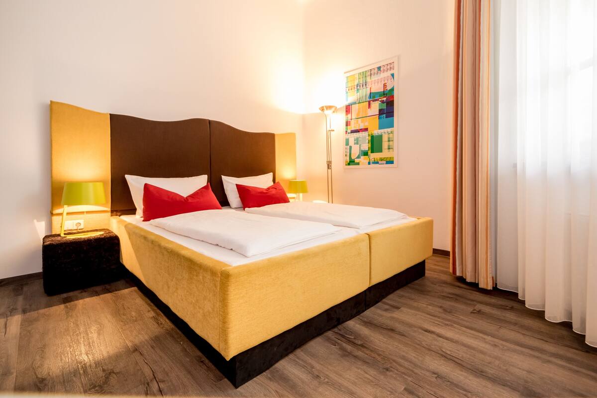 A hotel room with a large bed, two bedside lamps, a headboard, and a colorful abstract painting on the wall.
