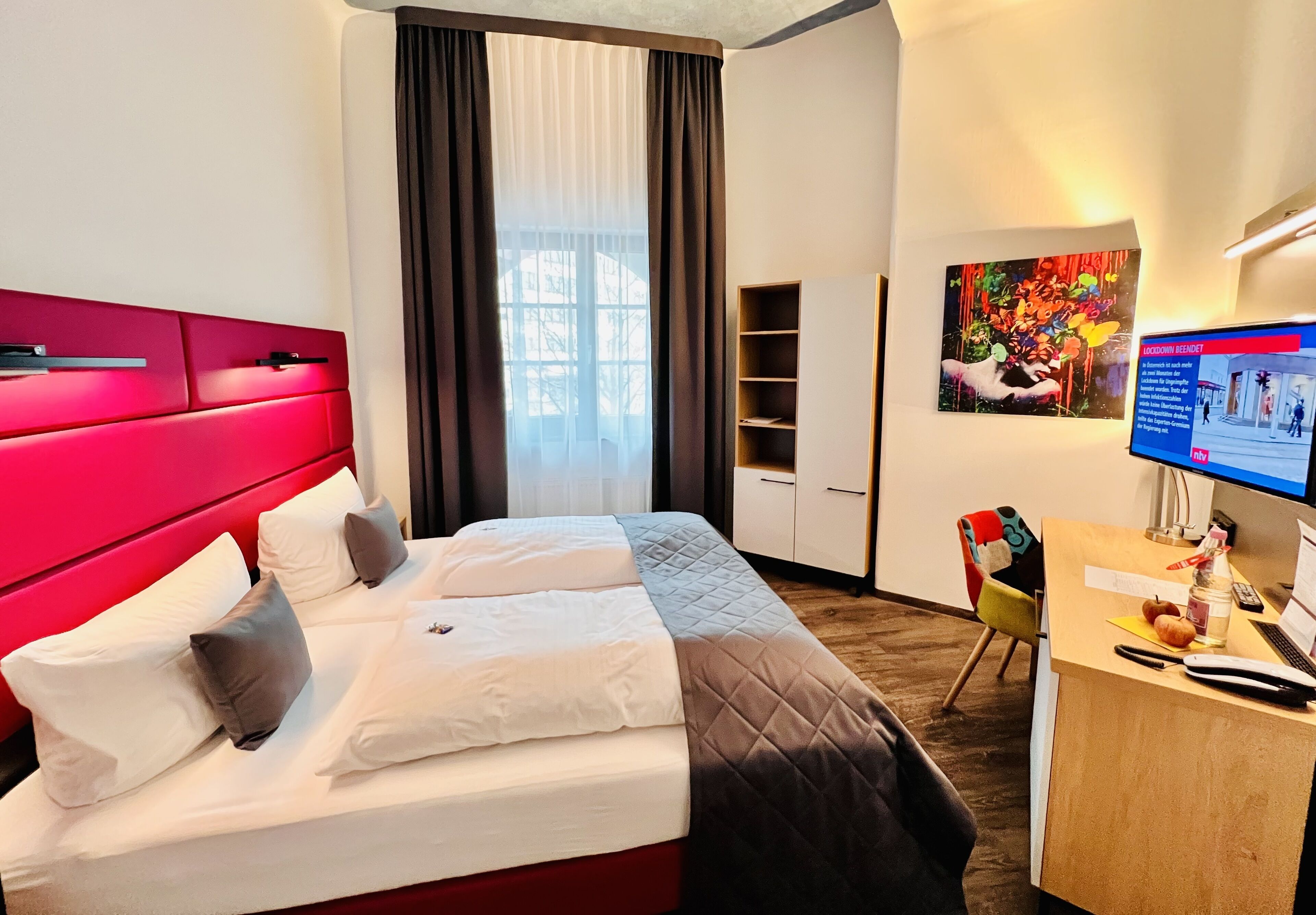 A hotel room with a red headboard, a bed with white and brown bedding, a desk with a computer, and a window with curtains.