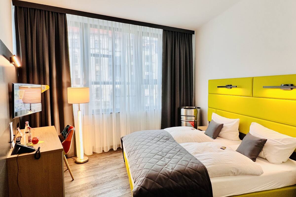 A hotel room with a bed, a desk with a computer, a lamp, and a window with curtains.