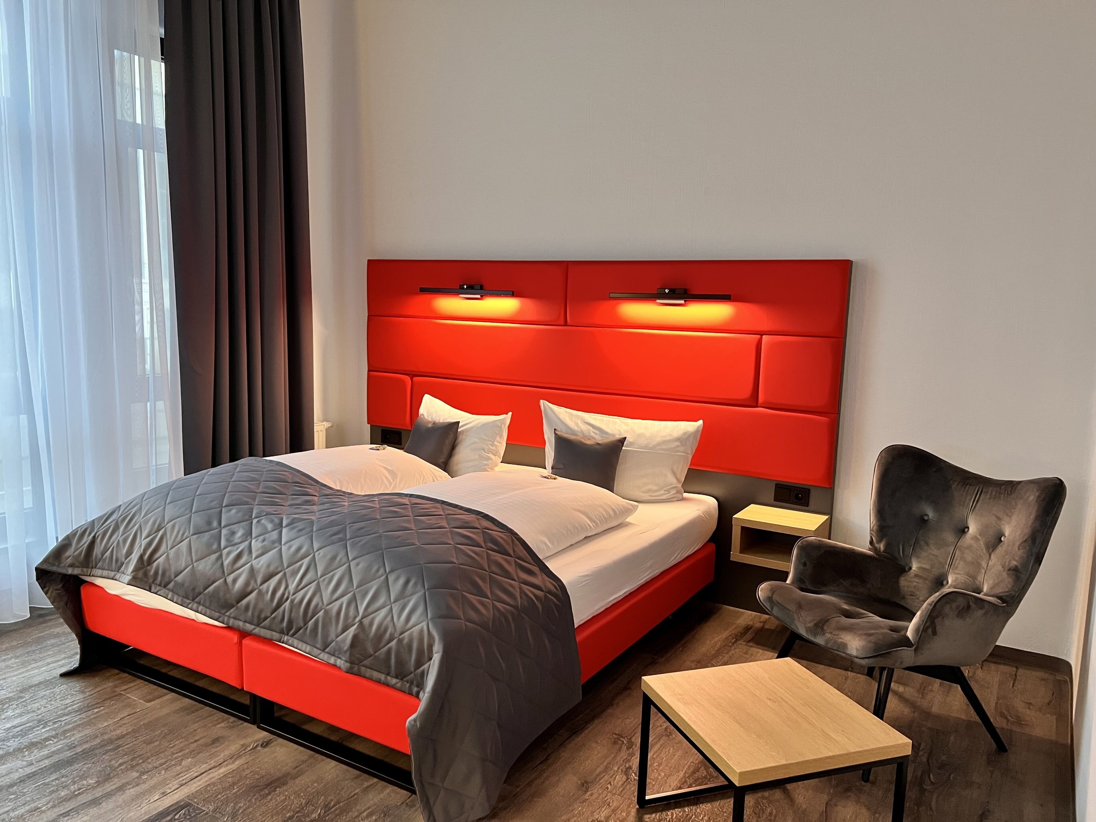 A modern hotel room with a red headboard, a bed with a quilted cover, a wooden nightstand, a chair, and a small table.