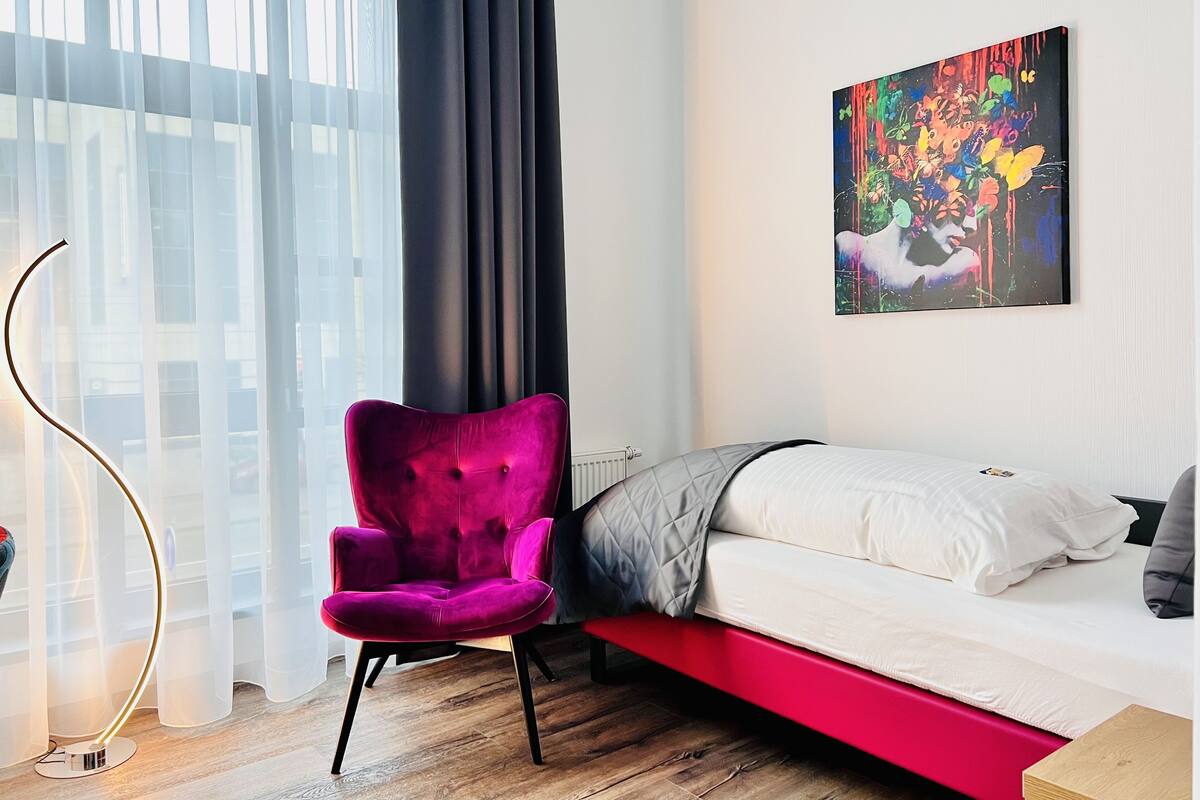 A modern bedroom with a bed, a vibrant purple chair, a floor lamp, and a colorful abstract painting on the wall.