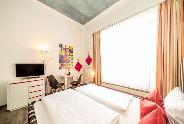 A hotel room with two beds, a desk with a TV, a chair, and a window with curtains. - artHotel Magdeburg (Magdeburg)