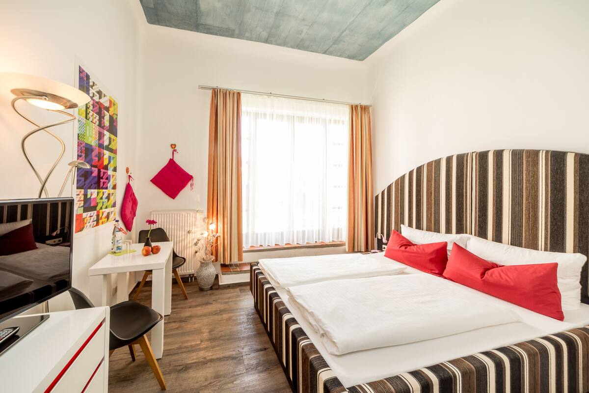 A hotel room with a bed, a desk, a chair, a lamp, and a window with curtains.