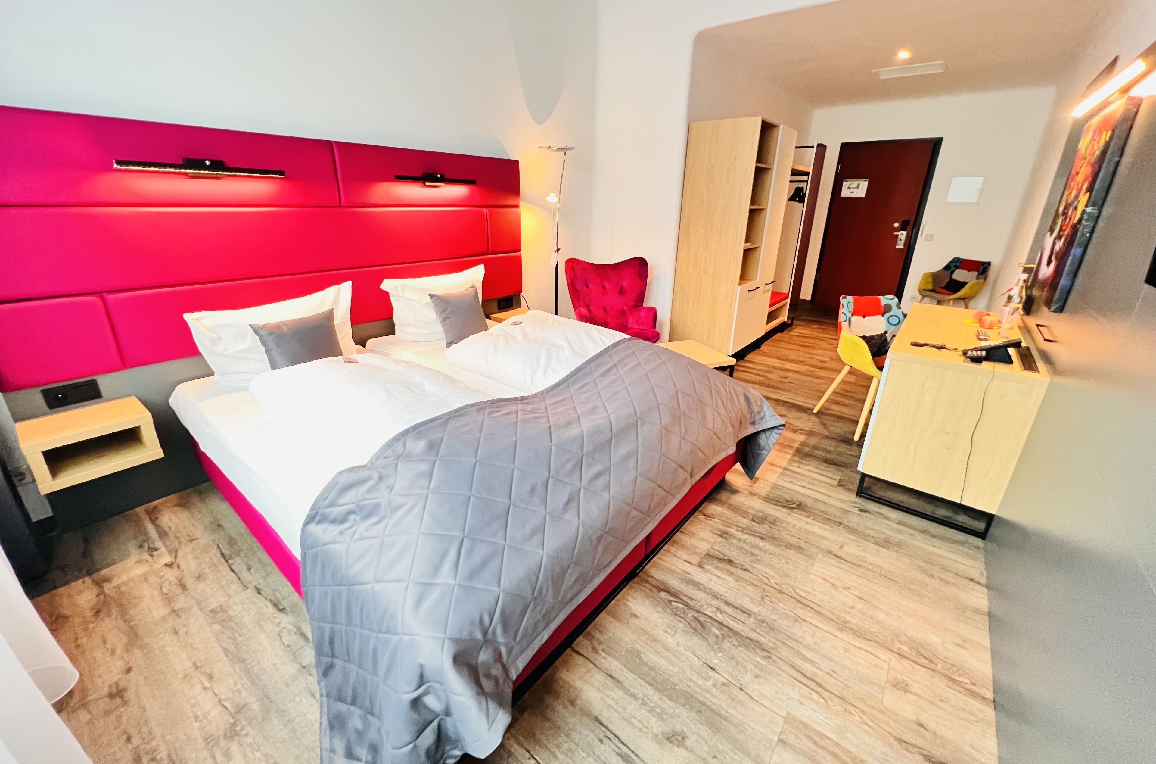 A modern hotel room with a large bed, a red headboard, a wooden wardrobe, and a small desk with a chair.