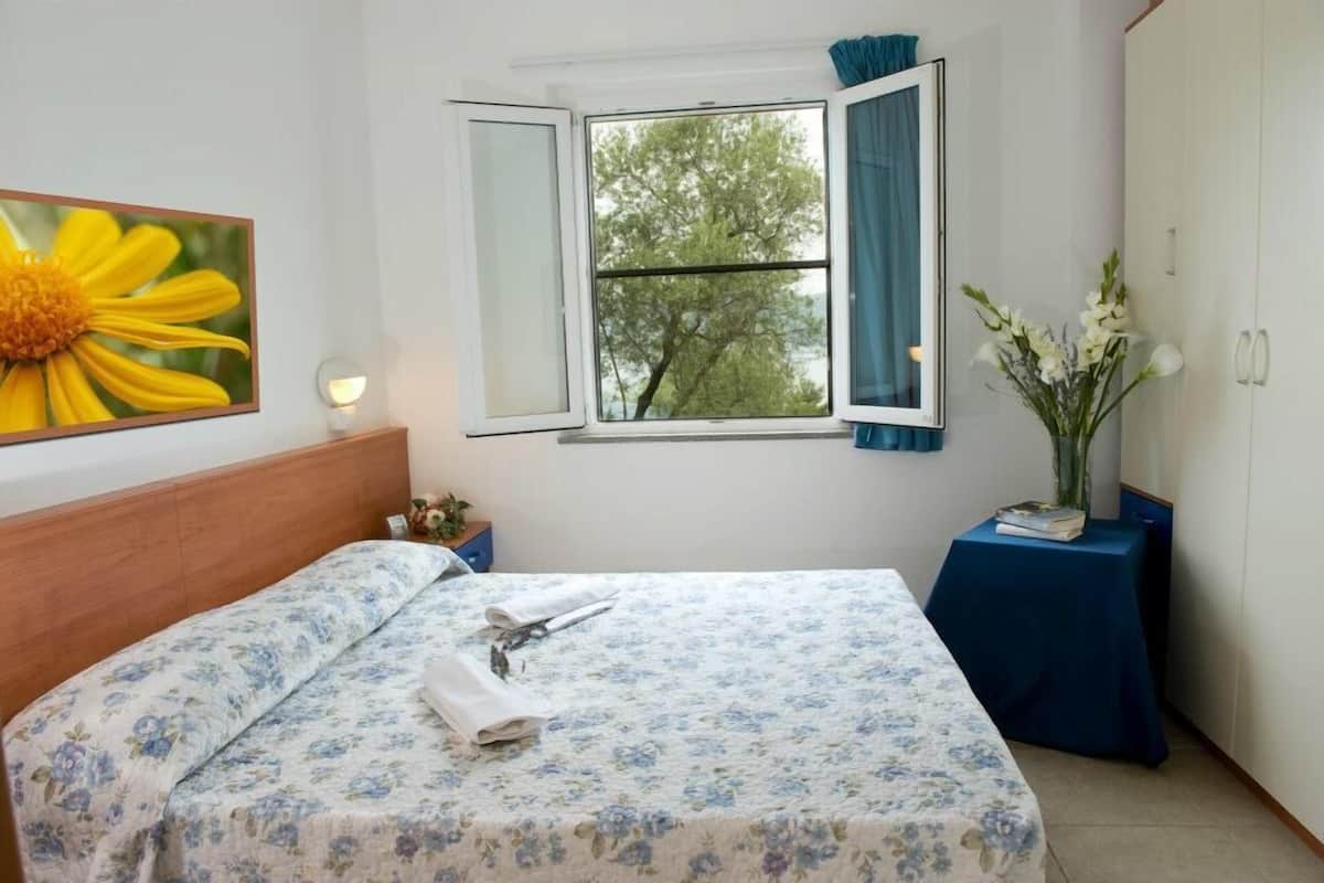Ein Hotelzimmer mit Bett, einem Fenster mit Blick auf Bäume, einem Blumenbild und einer Blumenvase.