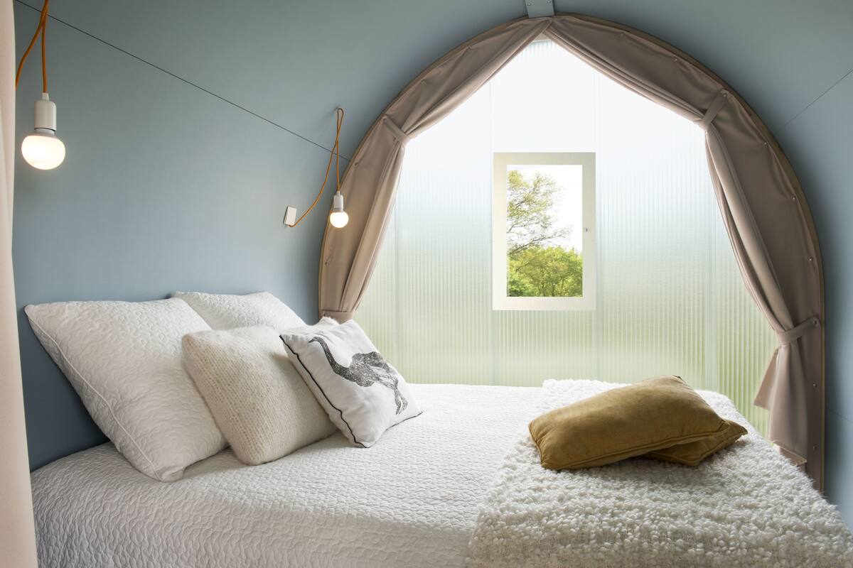 Ein Schlafzimmer mit Bett, Kissen, einem Fenster und Blick auf Grün.