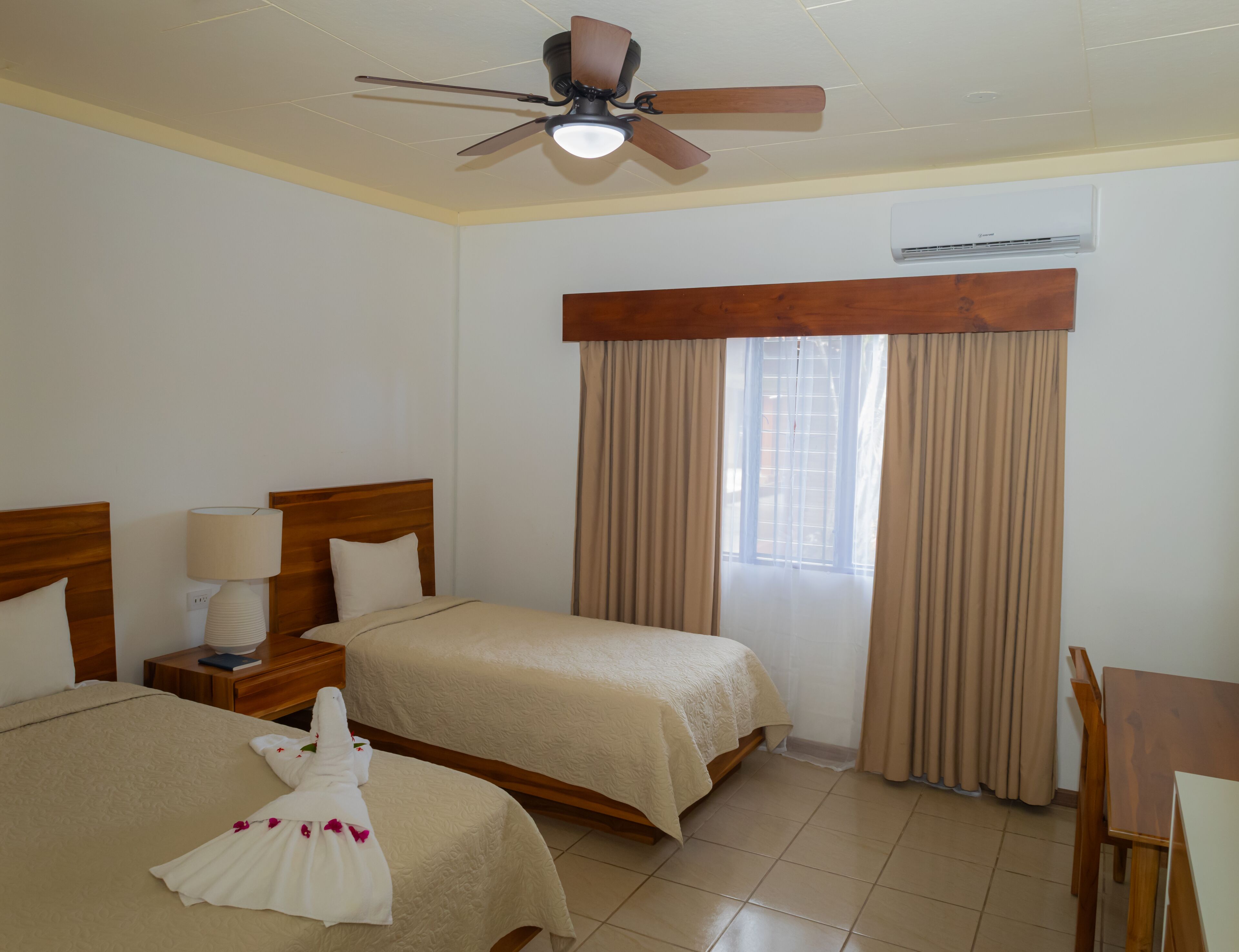 Habitación de hotel con dos camas, ventilador de techo, unidad de aire acondicionado y ventana con cortinas.