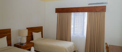 Habitación de hotel con dos camas, ventilador de techo, unidad de aire acondicionado y ventana con cortinas.