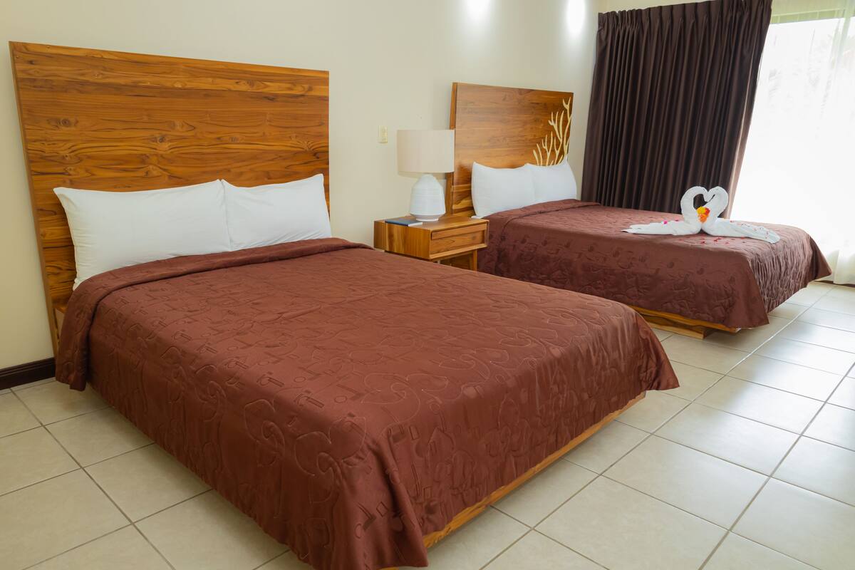 Two beds with brown bedding, wooden headboards, a nightstand with a lamp, and a window with curtains.