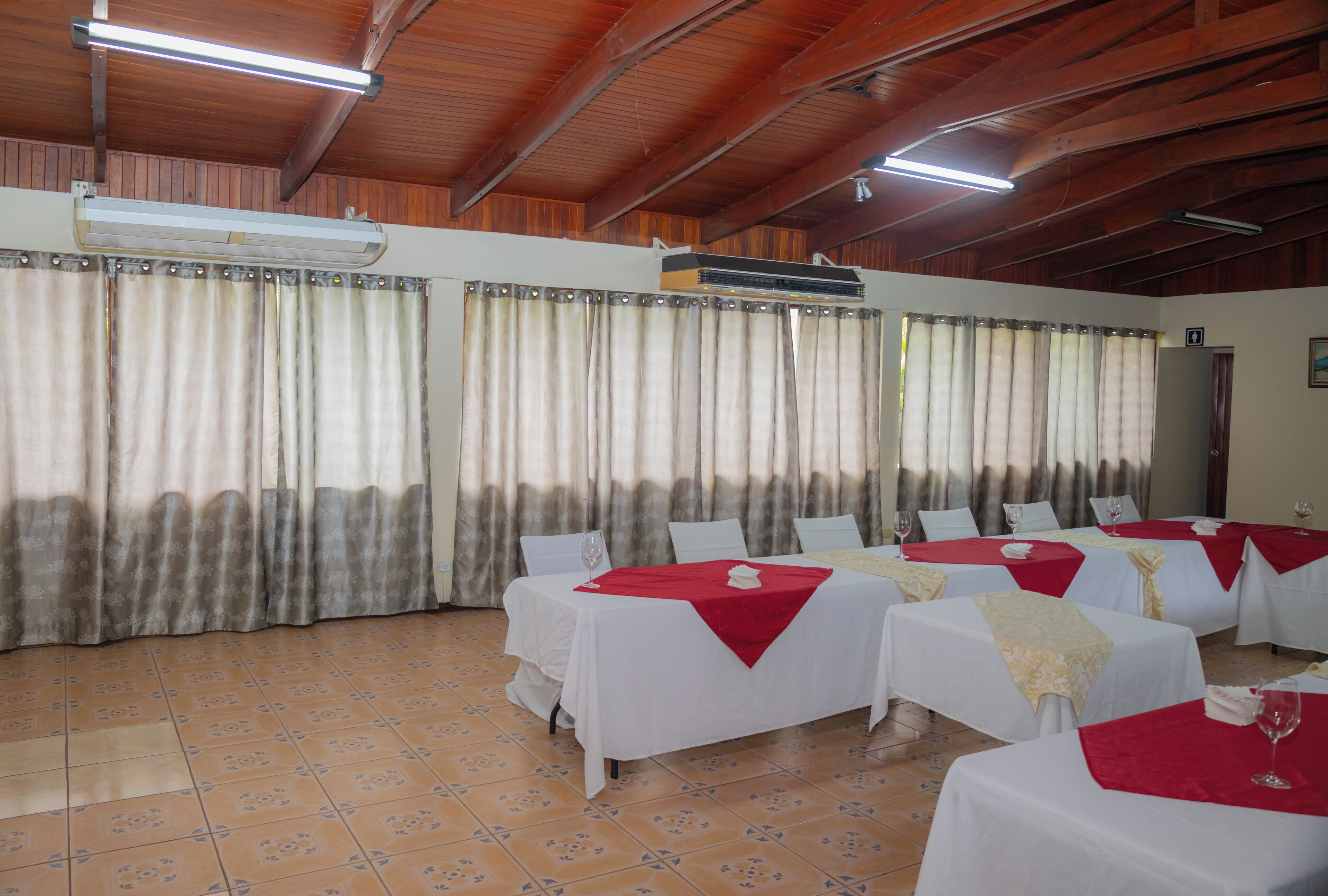 Un salón de banquetes con varias mesas dispuestas para comer, unidades de aire acondicionado y cortinas en las ventanas.