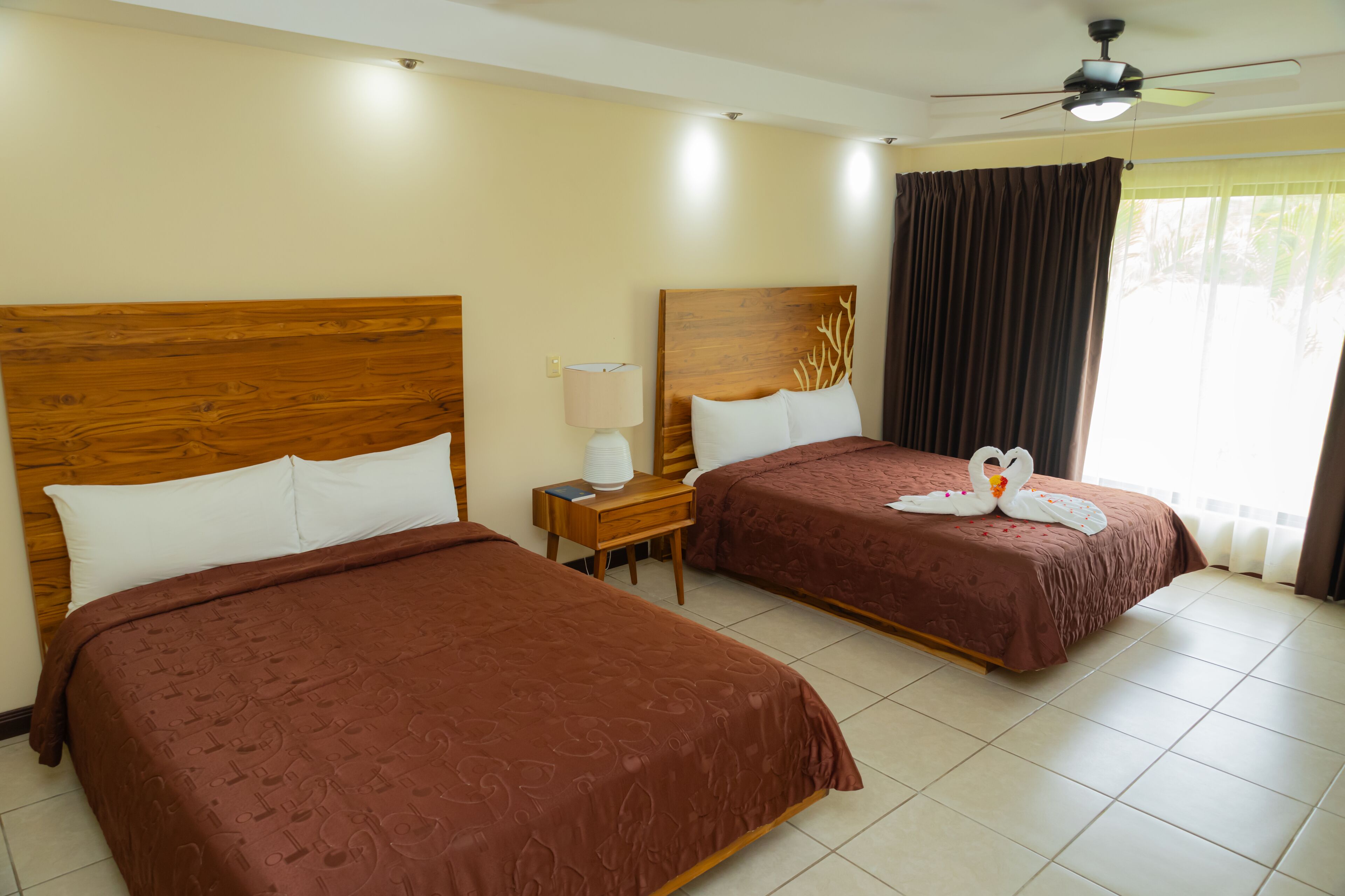 Une chambre d’hôtel avec deux lits, un ventilateur de plafond, une table de chevet avec une lampe et une fenêtre avec des rideaux.