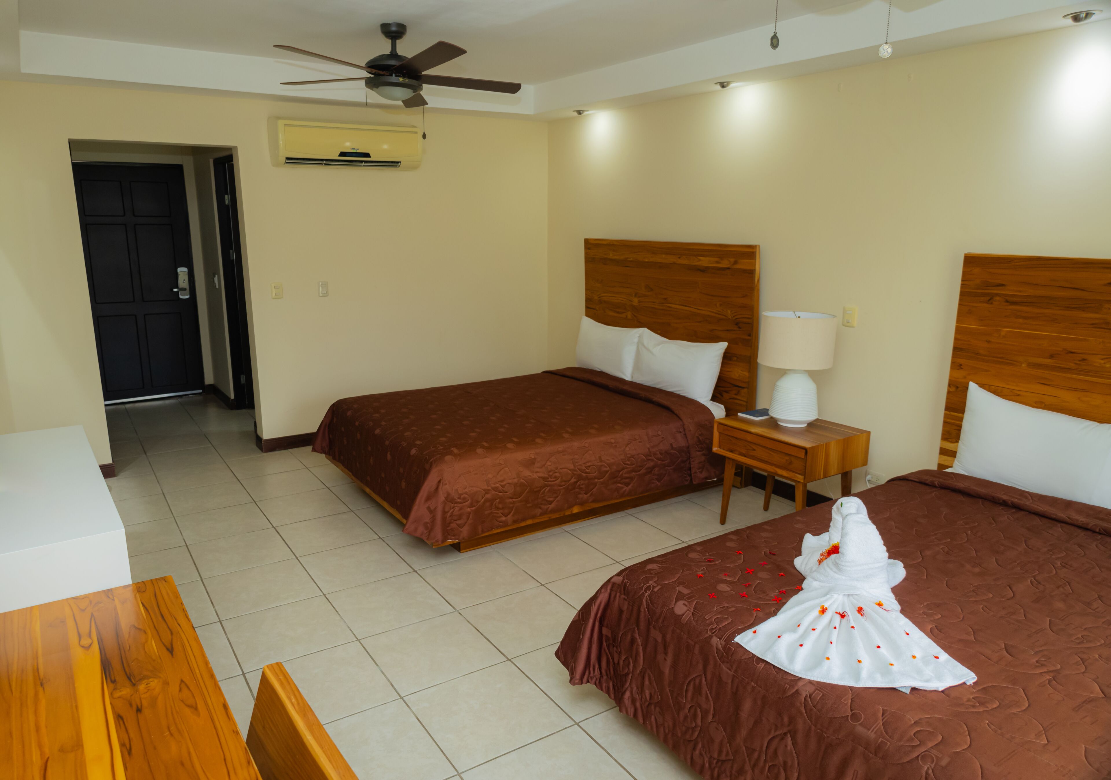 A hotel room with two beds, a ceiling fan, a bedside table with a lamp, and a wooden desk.