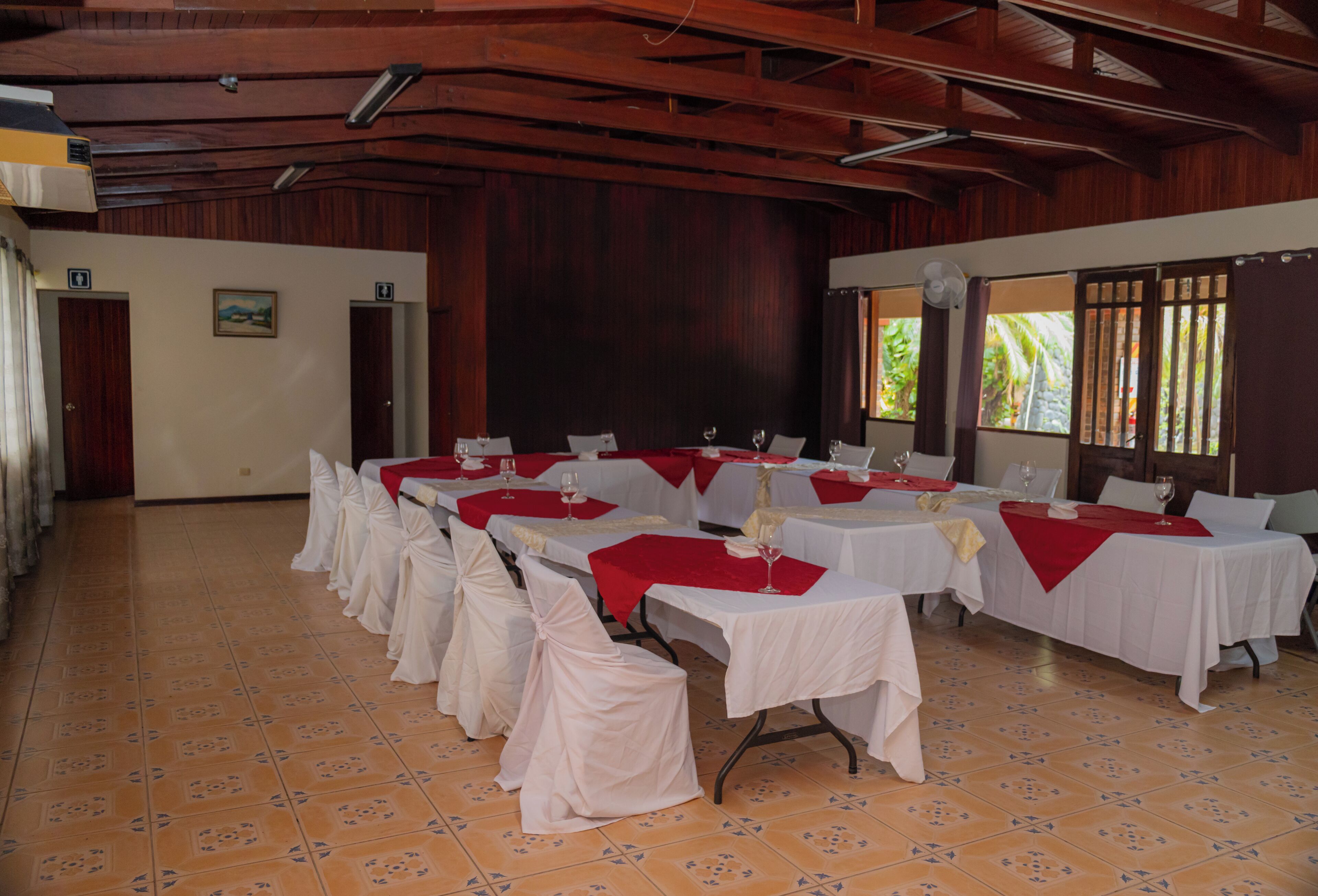 A banquet hall with multiple tables set for dining, wooden ceiling beams, and a view of greenery outside.