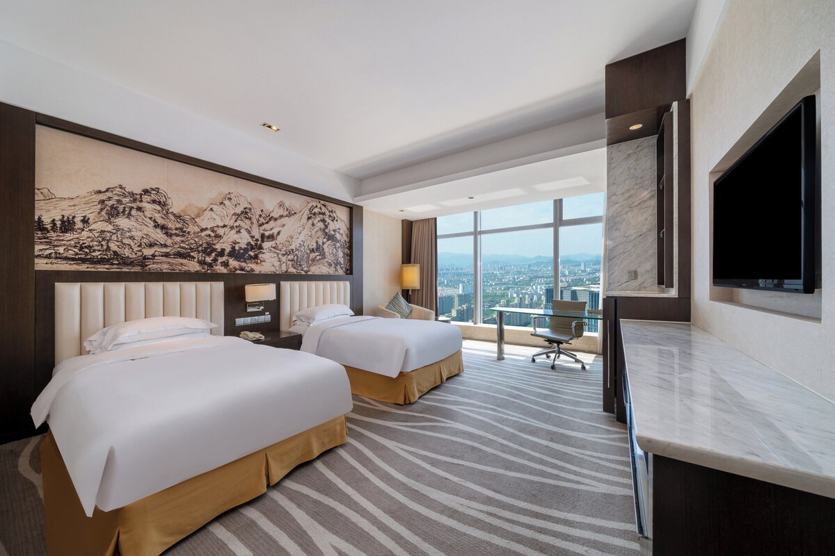 A hotel room with two beds, a large painting, a desk, and a city view.