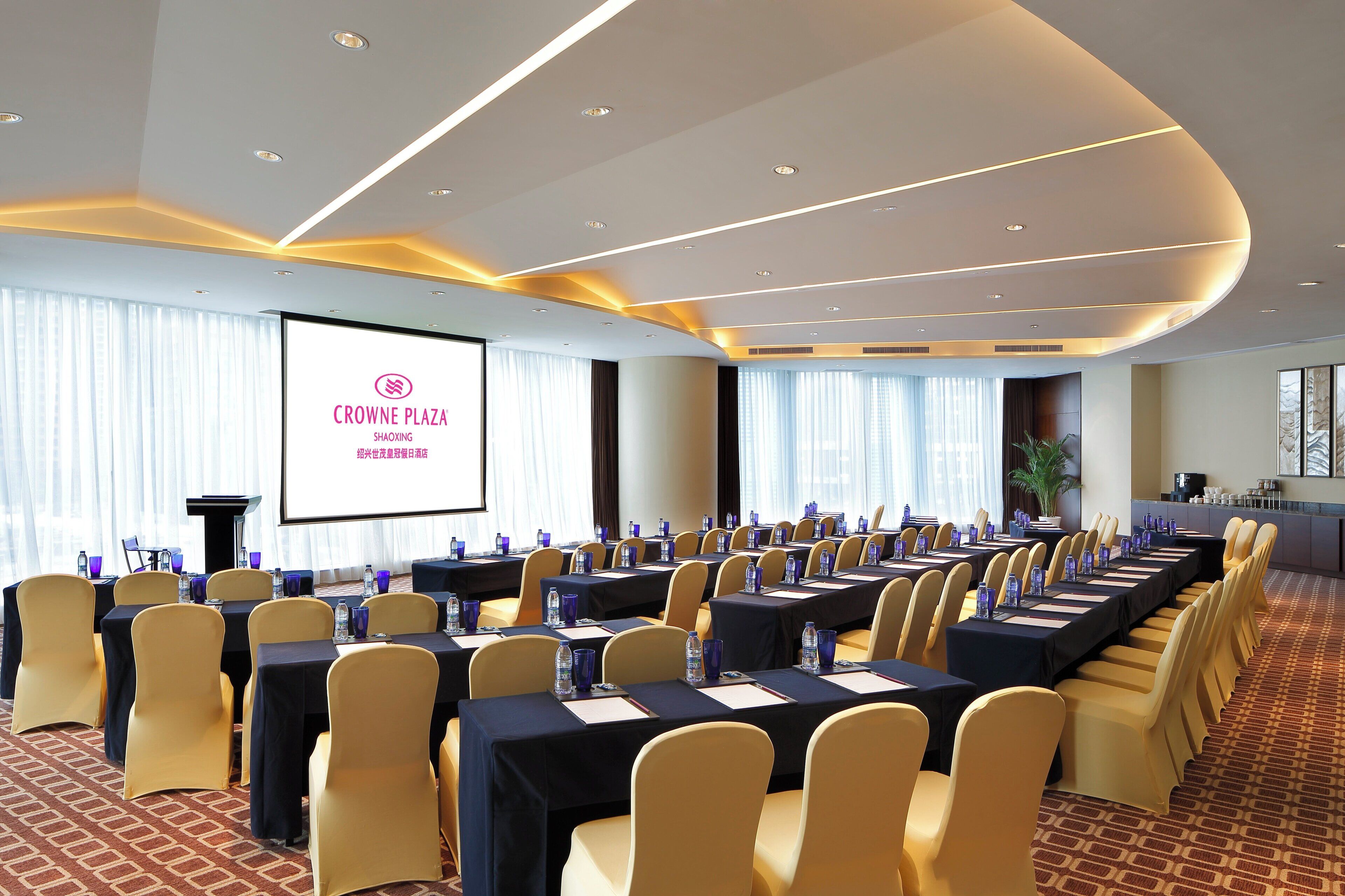 Een congreszaal met rijen tafels en stoelen, een scherm waarop 'CROWNE PLAZA SHAOXING' staat en een groot raam met gordijnen.