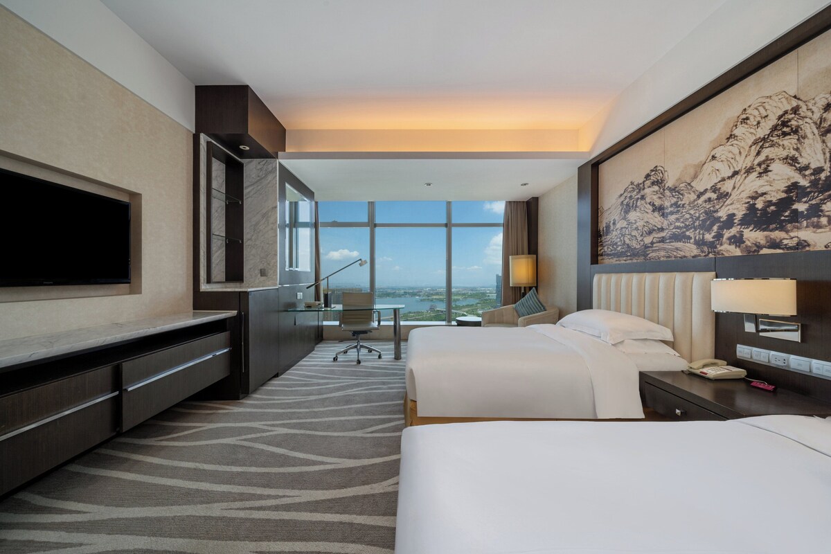 A hotel room with a large bed, a desk, a chair, a TV, and a view of the city.