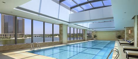 A large indoor swimming pool with clear views of a river and buildings, surrounded by sunlit areas with seating.