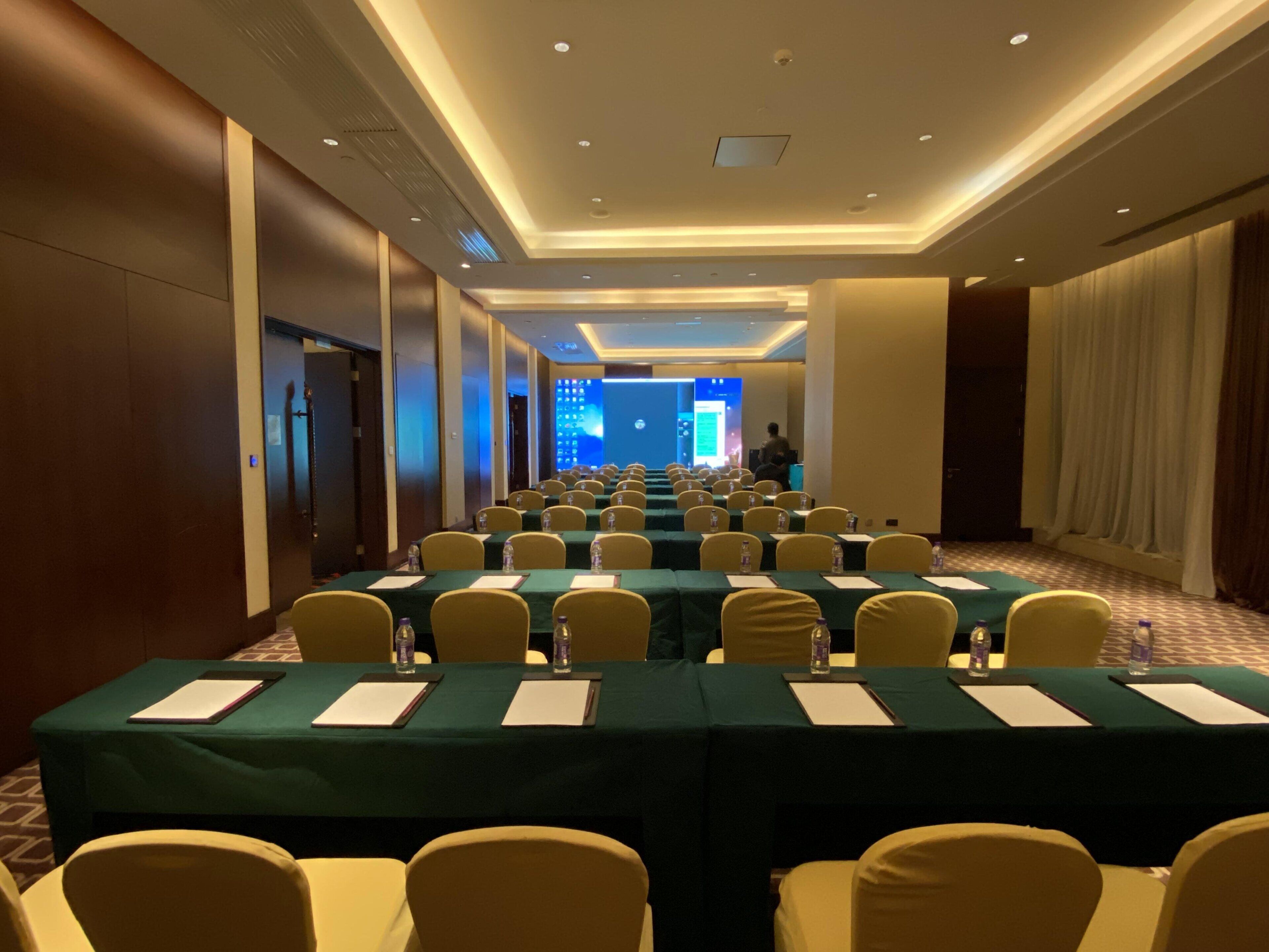 A conference room with rows of tables, chairs, and a presentation screen at the front.