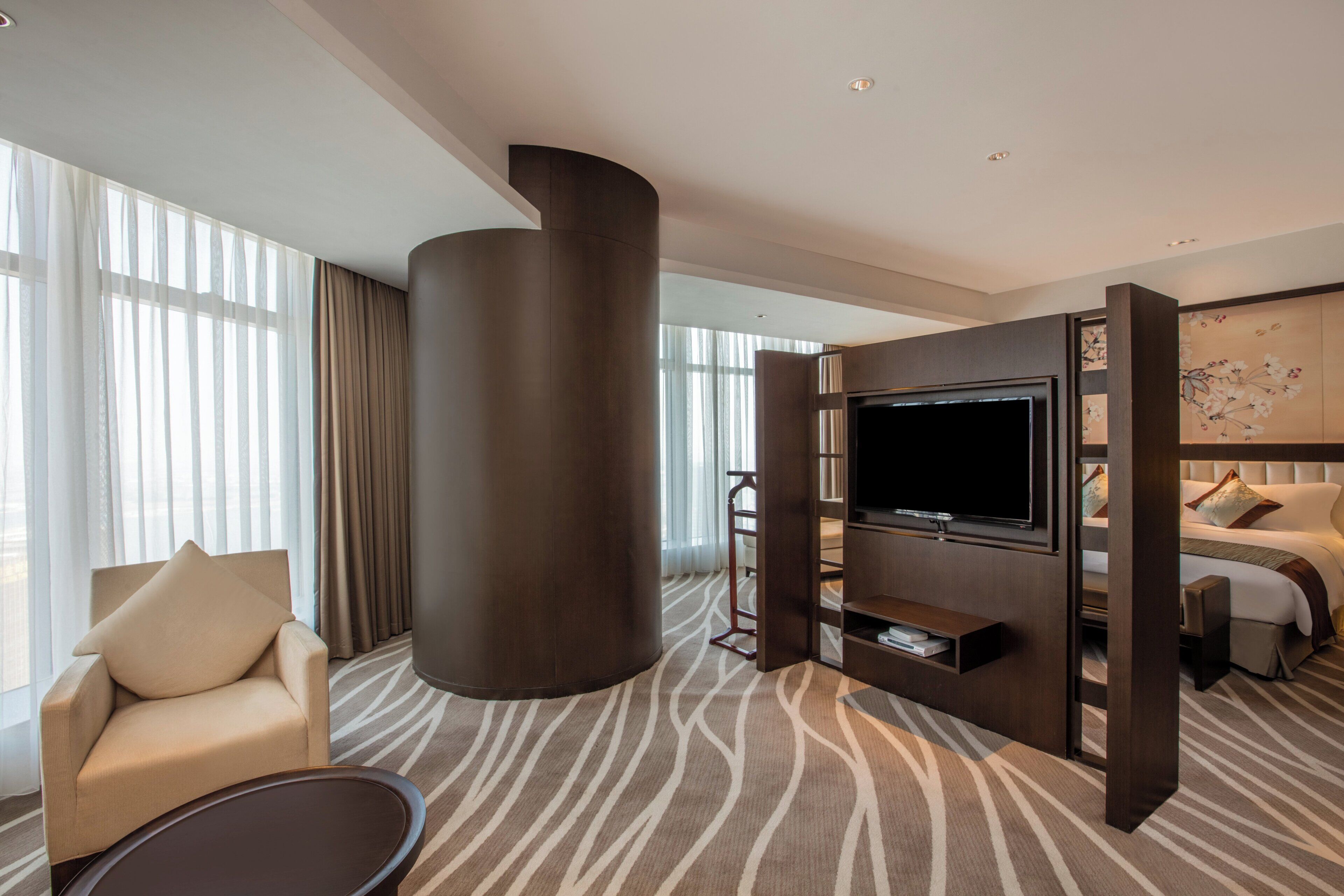 A modern hotel room with a large flat-screen TV, a sofa, a chair, and a decorative wall panel.