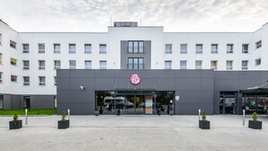 A modern building with a prominent 'fh' logo on the facade, large windows, and a covered entrance area with potted plants. - Focus Hotel Katowice Chorzow (Chorzow)