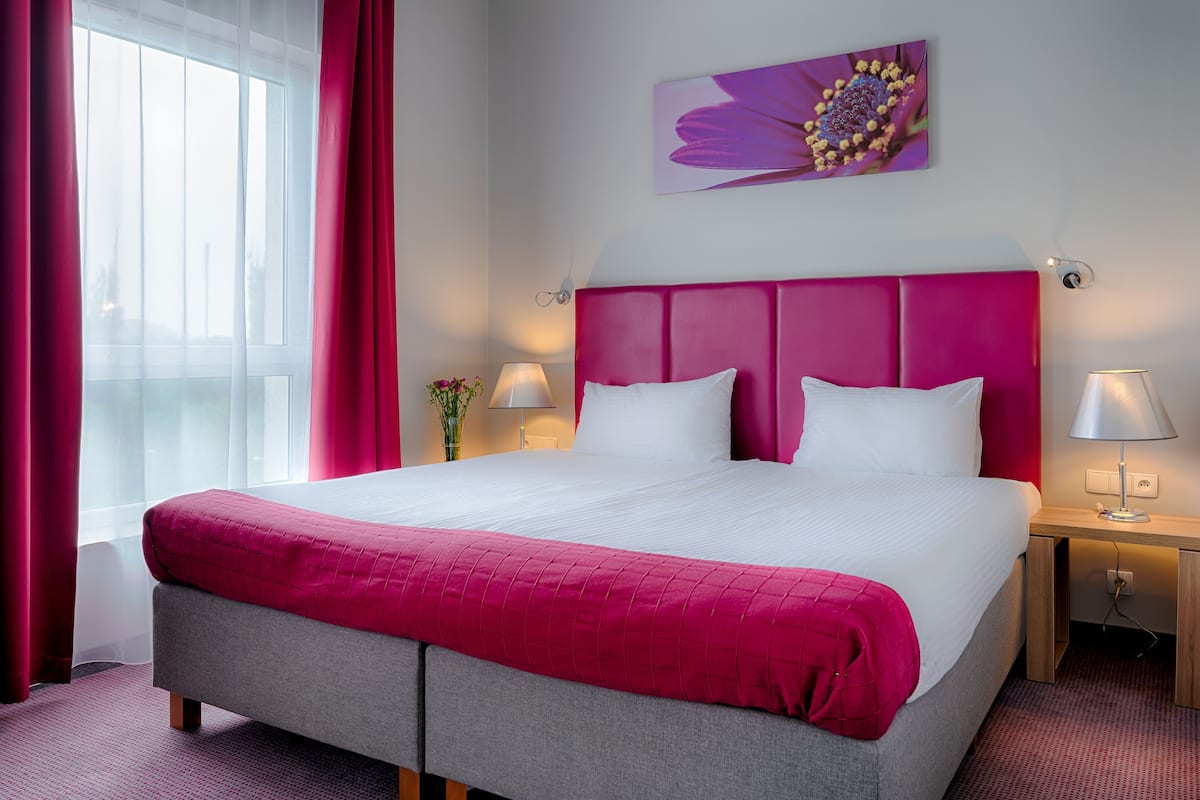Une chambre d’hôtel avec un grand lit, une tête de lit rose et une fenêtre avec des rideaux.
