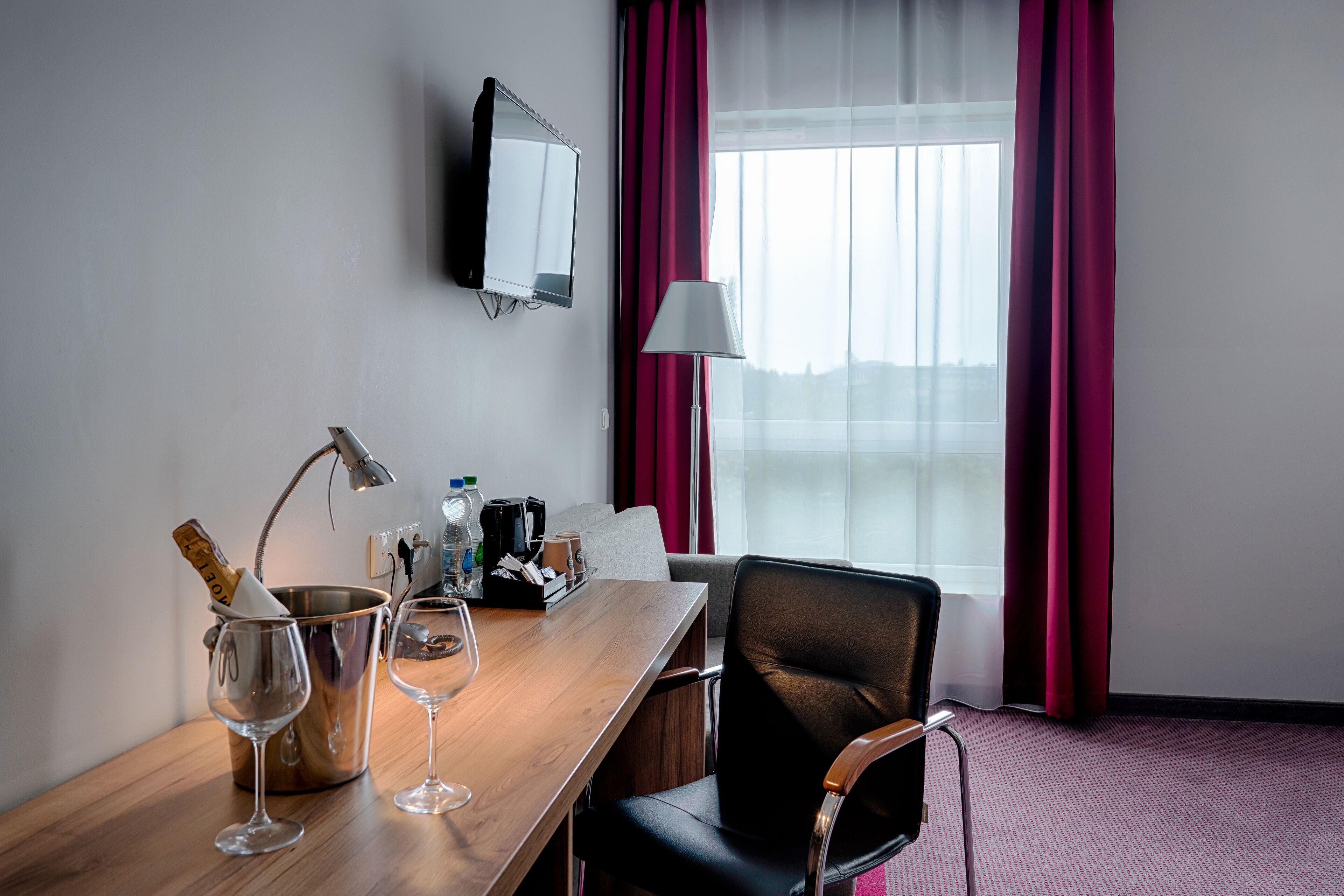 A hotel room with a wooden desk, two wine glasses, a bottle of champagne, a lamp, a TV, and a window with red curtains.