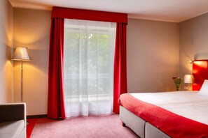 Une chambre d’hôtel avec un lit, un bureau, une lampe et une fenêtre avec des rideaux rouges.