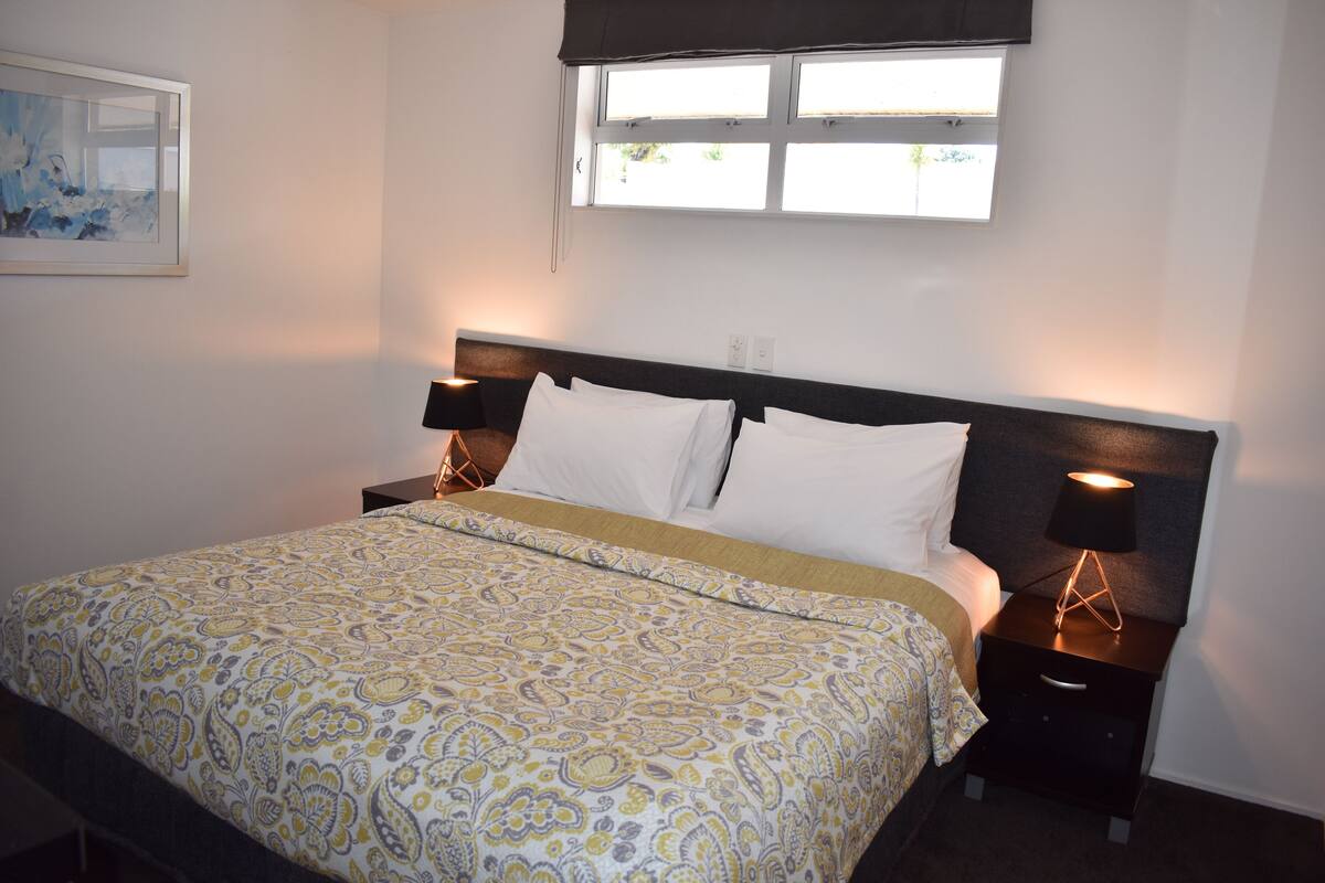 A neatly made bed with a patterned bedspread, a bedside lamp, and a window with a view of greenery.