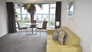 Una habitación con un sofá amarillo, dos sillones, una mesita redonda y un gran ventanal con cortinas.