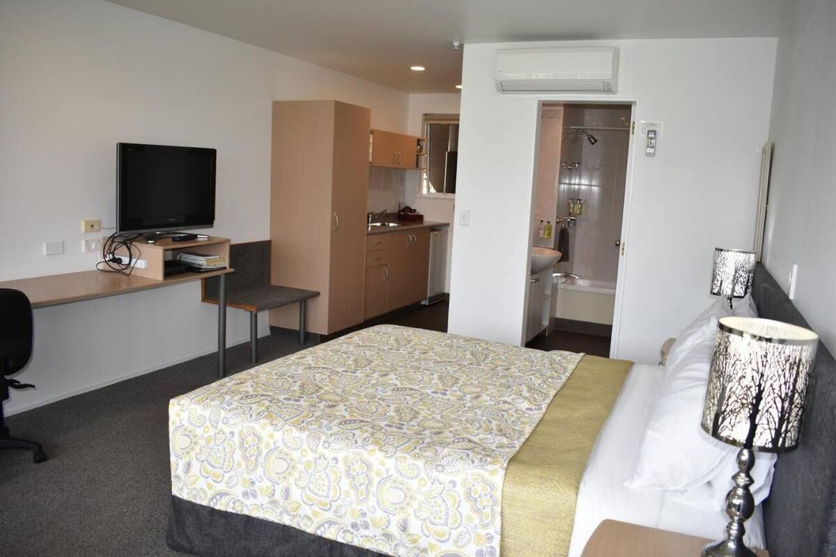 A hotel room with a bed, a desk with a TV, a kitchenette, and a bathroom.