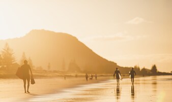 Uma praia com pessoas caminhando e carregando pranchas de surf, com uma montanha ao fundo.