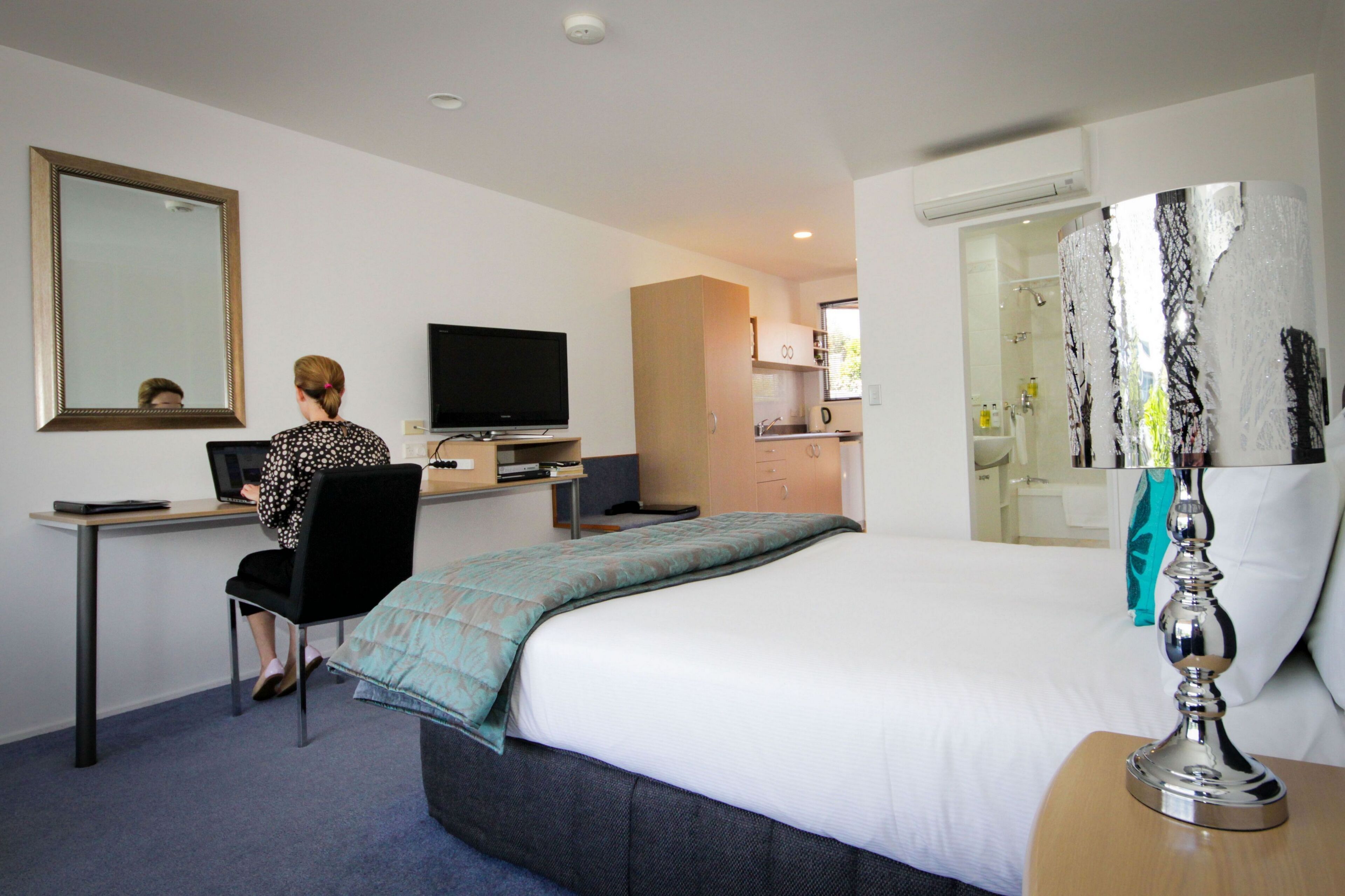 A hotel room with a bed, a desk with a computer, a television, and a bathroom visible through an open door.