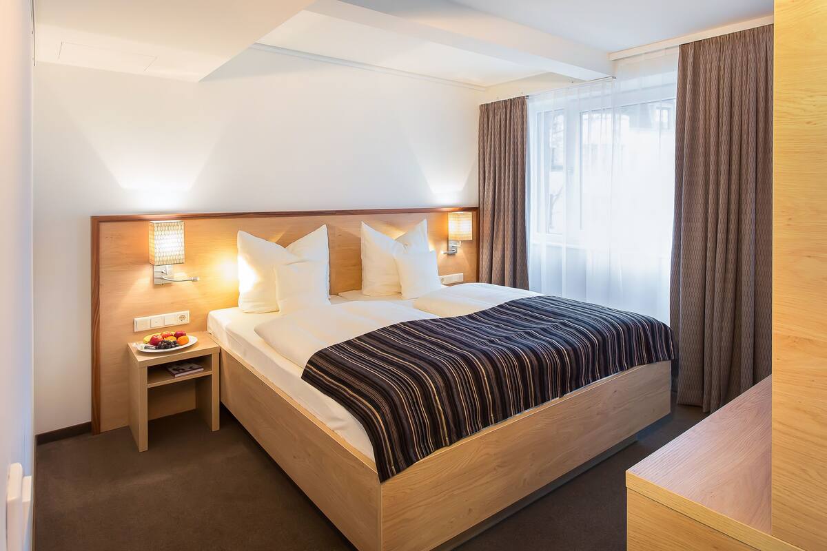 Ein Hotelzimmer mit einem hölzernen Bett, einem Nachttisch mit Obst, einer Lampe und Blick ins Freie.