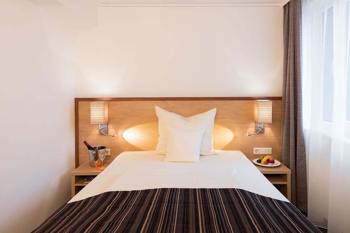 Ein Hotelzimmer mit einem großen Bett, zwei Nachttischlampen, einem Nachttisch mit Obstteller und einem Fenster mit Vorhängen.
