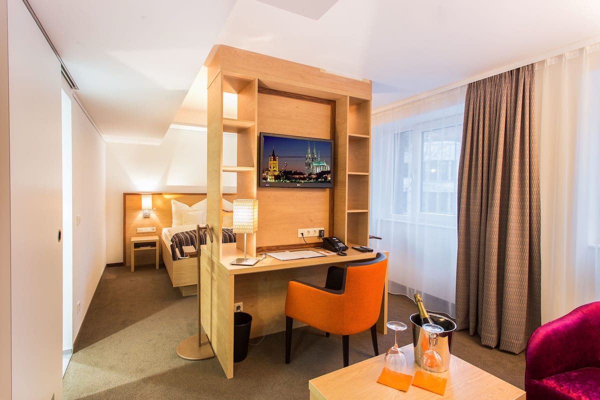 Ein Hotelzimmer mit Holzschreibtisch, Fernseher, Bett, Sessel, einem kleinen Tisch mit Flasche und Glas sowie einem roten Sessel.