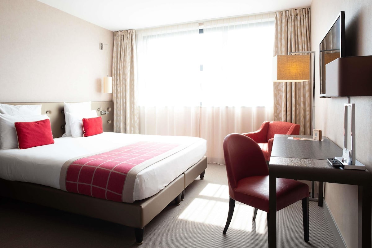 Habitación de hotel con cama, escritorio, silla, televisor y ventana con cortinas.