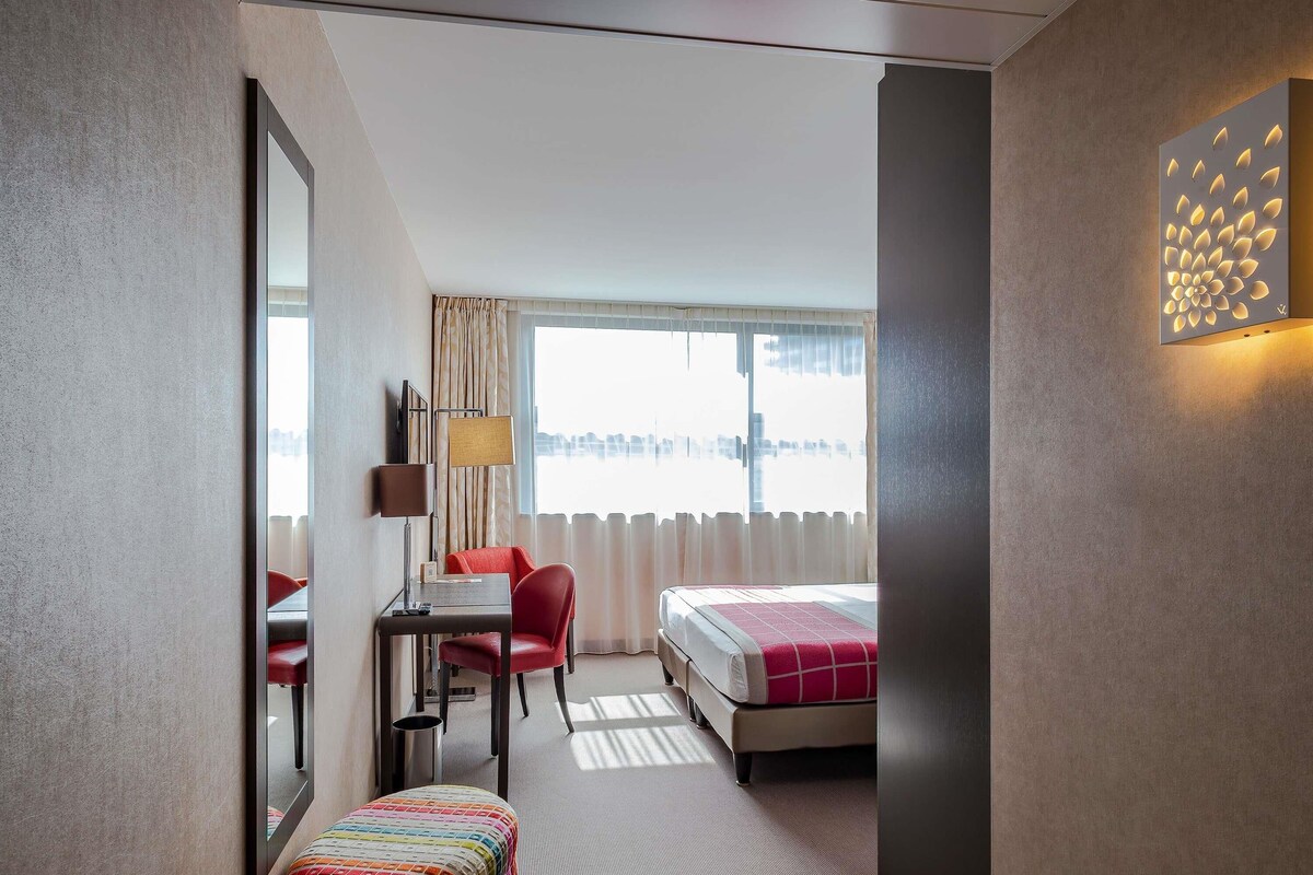 Hotelkamer met een bed, een bureau met stoel, een klein tafeltje met lamp, een kleurrijk pouf en een grote spiegel.