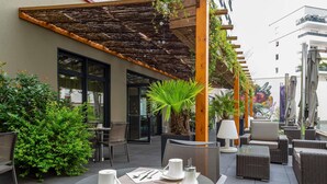 Un café al aire libre con mesas y sillas, plantas en macetas y una pérgola de madera.
