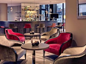 Zona de bar con diversas opciones de asientos, incluyendo sillas tapizadas en rojo y beige, una mesa redonda con objetos decorativos y una barra llena de diferentes botellas.