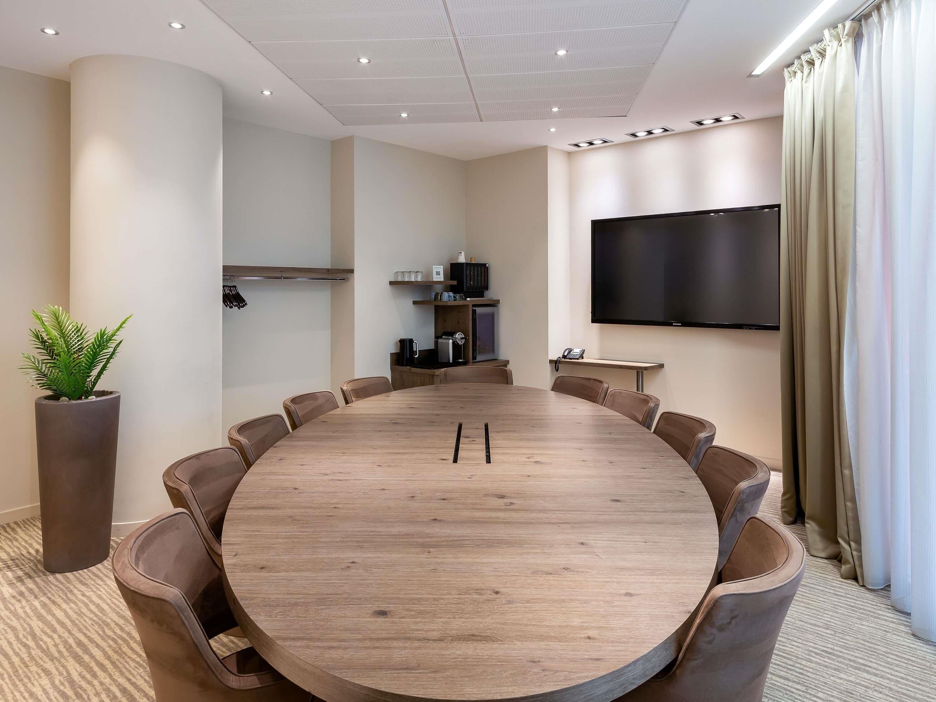 A conference room with a large round table, chairs, a TV screen, and a potted plant.
