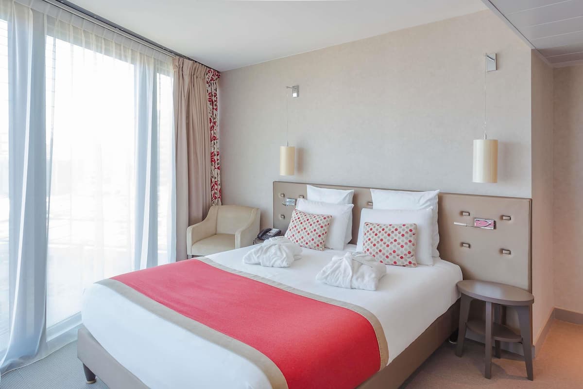 Habitación de hotel con una cama grande, una silla, una mesita de noche y vistas al exterior a través de la ventana.