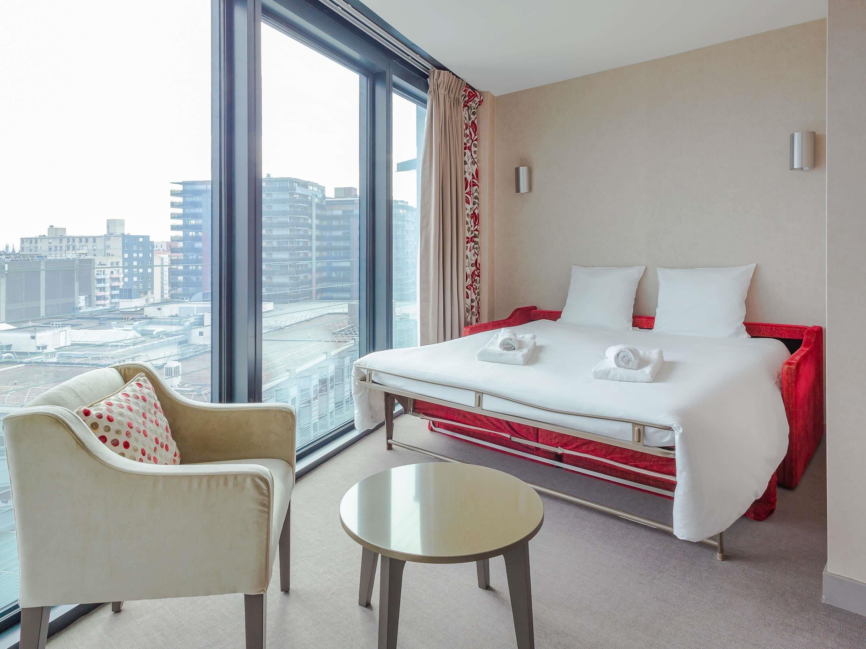 Quarto de hotel com uma cama, uma cadeira, uma mesa pequena e uma grande janela com vista para a cidade.