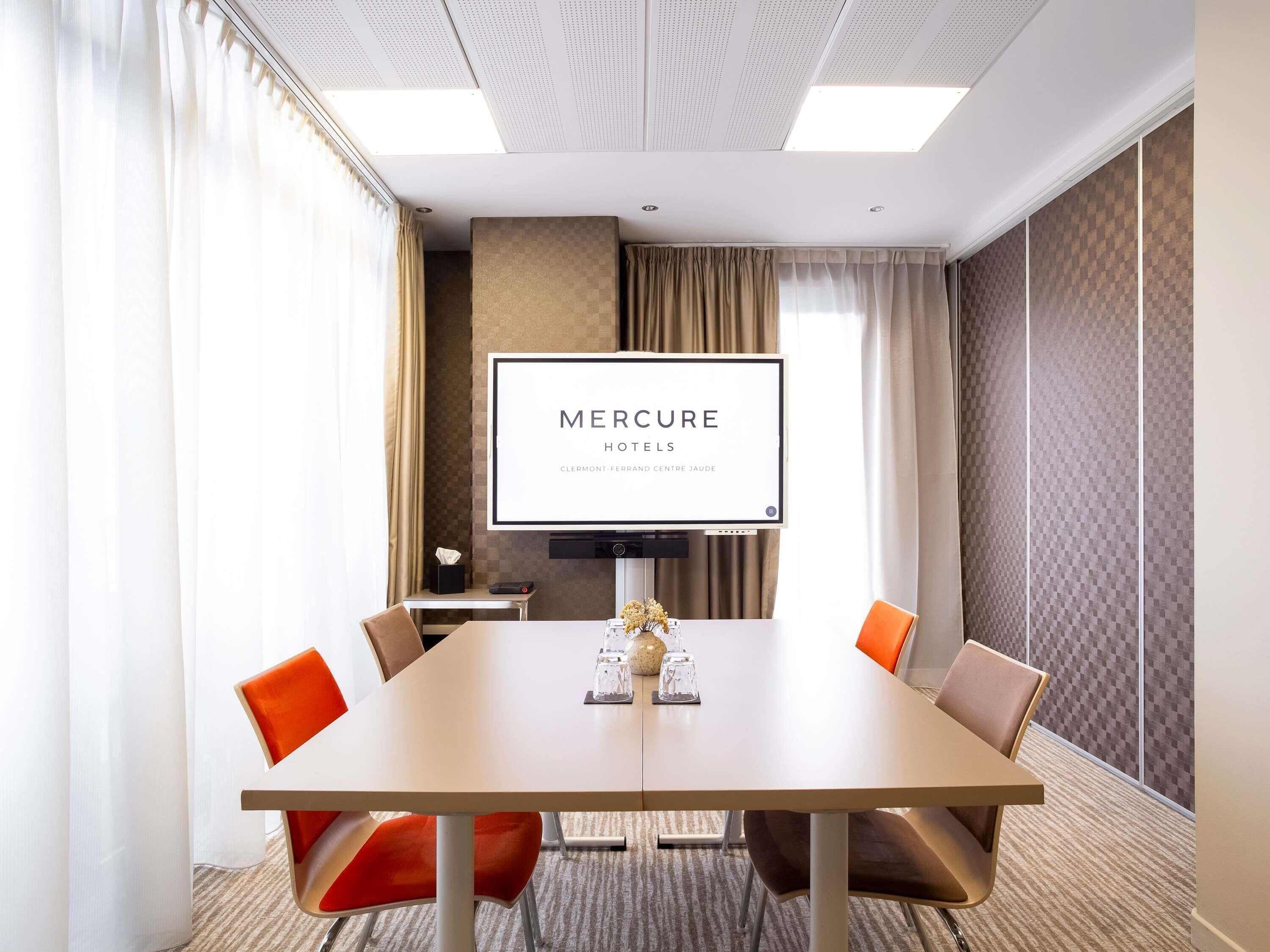 Un salón de conferencias con una mesa grande, cuatro sillas, una pantalla de televisión que muestra 'MERCURE HOTELS' y vistas de la ciudad a través de amplias ventanas.