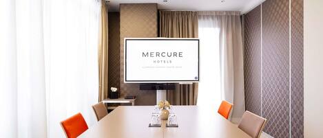 A conference room with a large table, four chairs, a television screen displaying 'MERCURE HOTELS,' and a view of the cityscape through large windows.