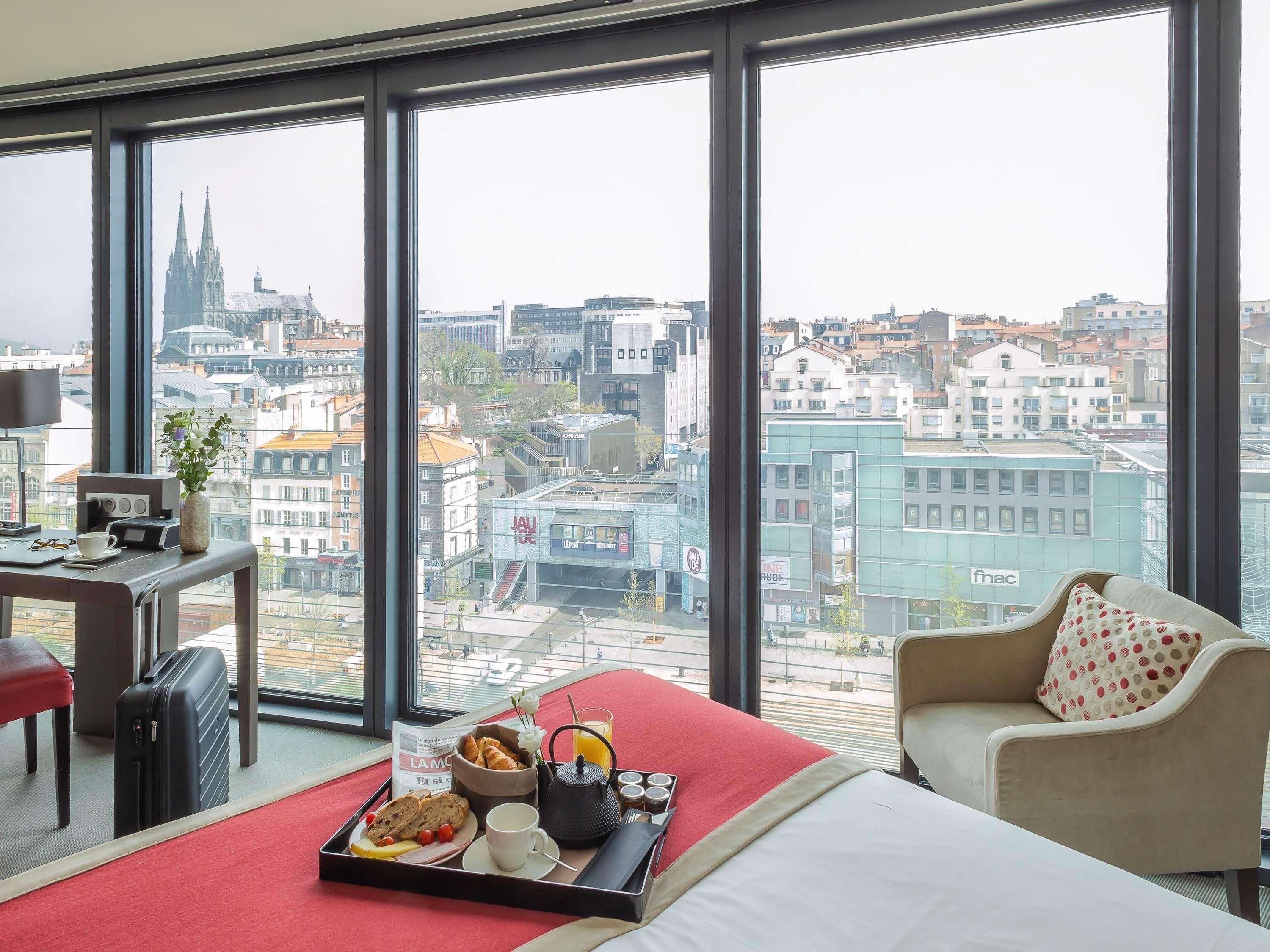 A hotel room with a large window, a bed, a desk, and a tray with breakfast items.