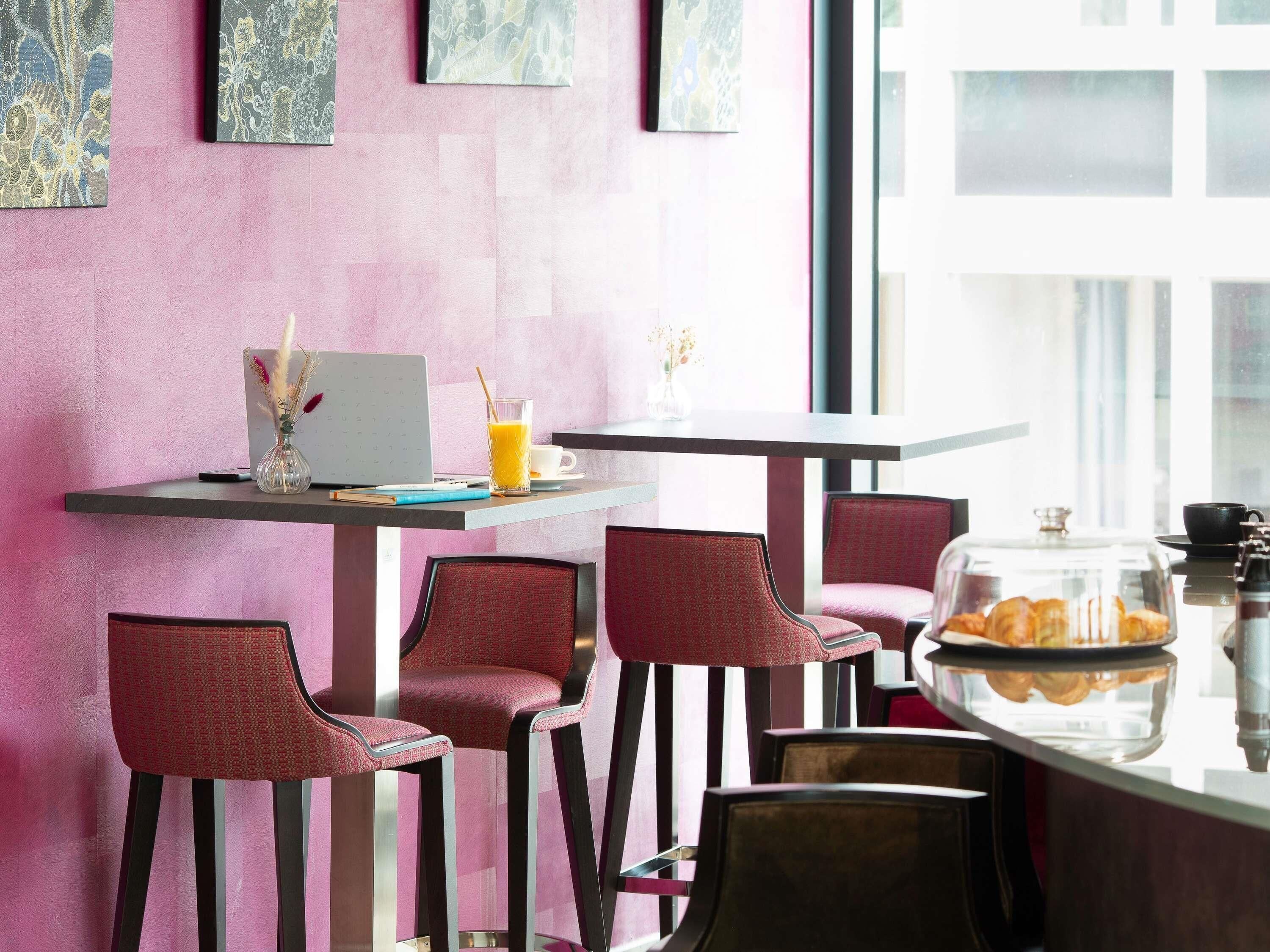 Un café moderne avec un mur rose, un ordinateur portable, un verre de jus d’orange et une vitrine remplie de pâtisseries.