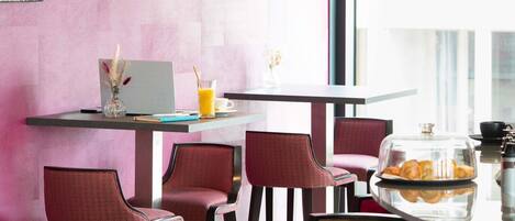 Un café moderne avec un mur rose, un ordinateur portable, un verre de jus d’orange et une vitrine remplie de pâtisseries.