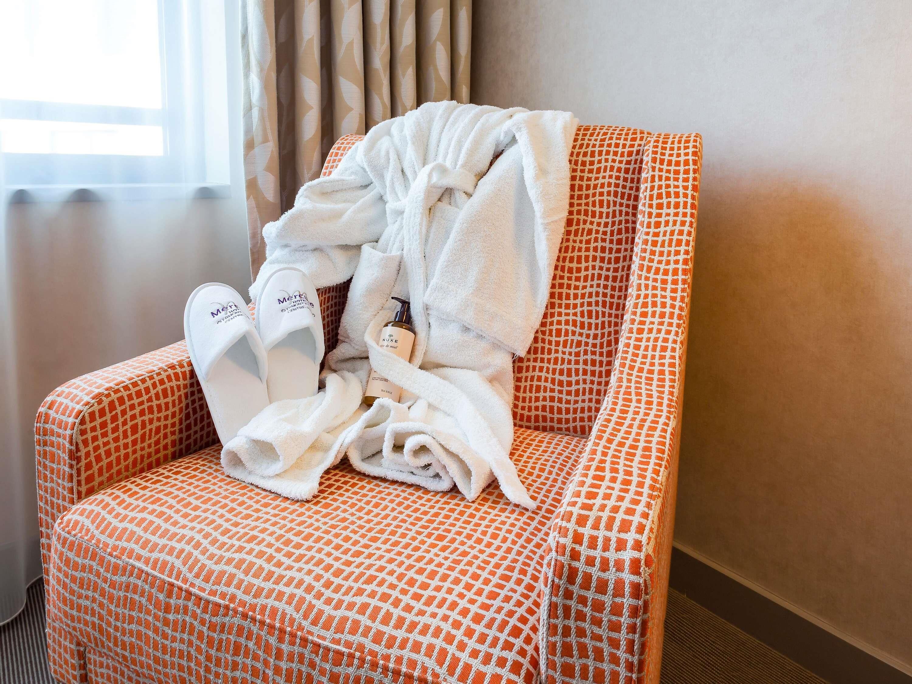A hotel room with a patterned orange and white chair, a white bathrobe, slippers, and a bottle of lotion.