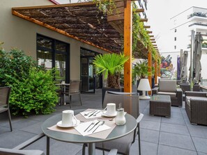 An outdoor patio with round tables, chairs, and umbrellas, surrounded by greenery and a building with a wooden pergola. - Mercure Clermont Ferrand Centre Jaude (Clermont-Ferrand)