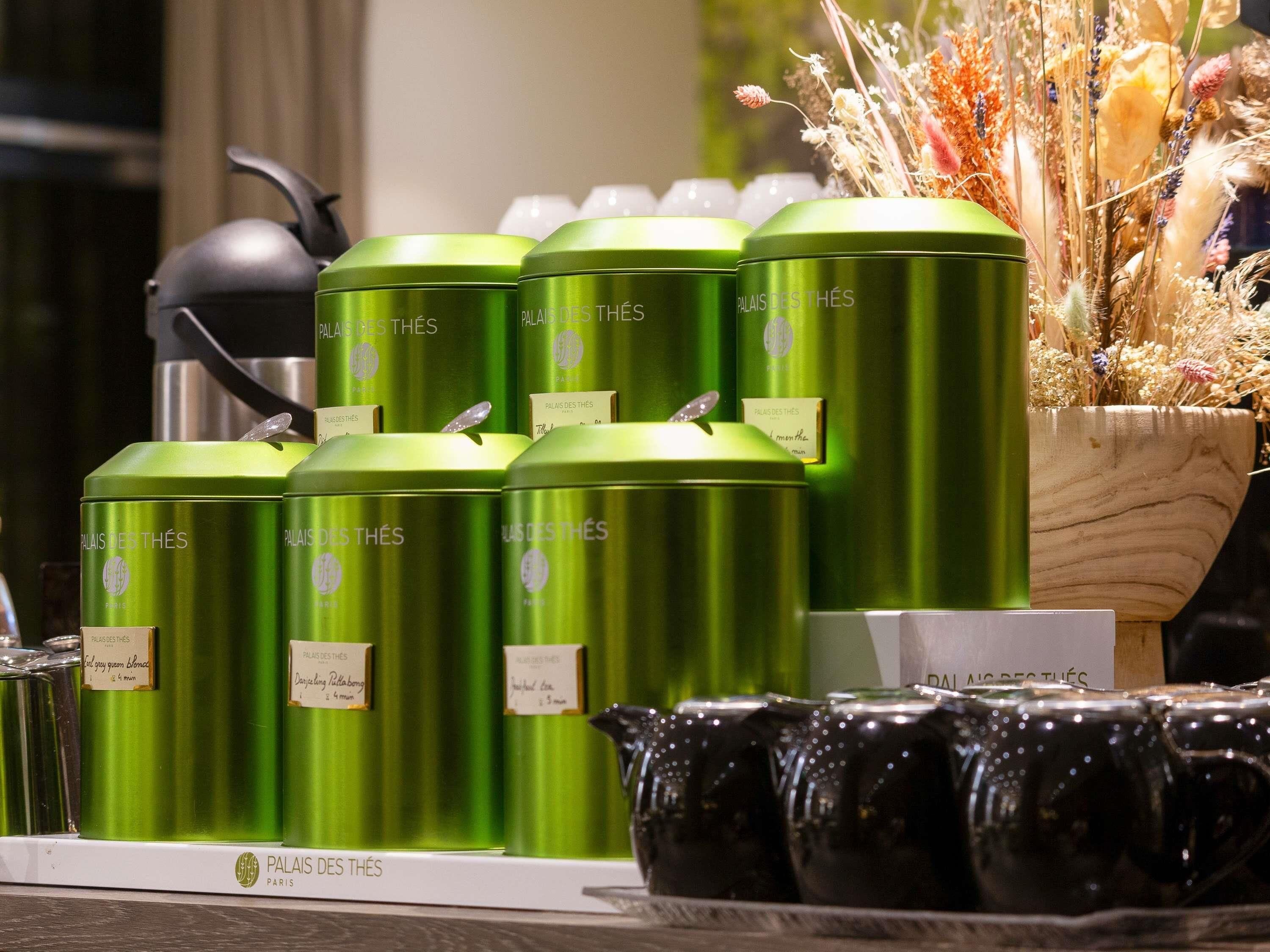 A display of green metal containers with labels, a black teapot, and black ceramic cups on a shelf.